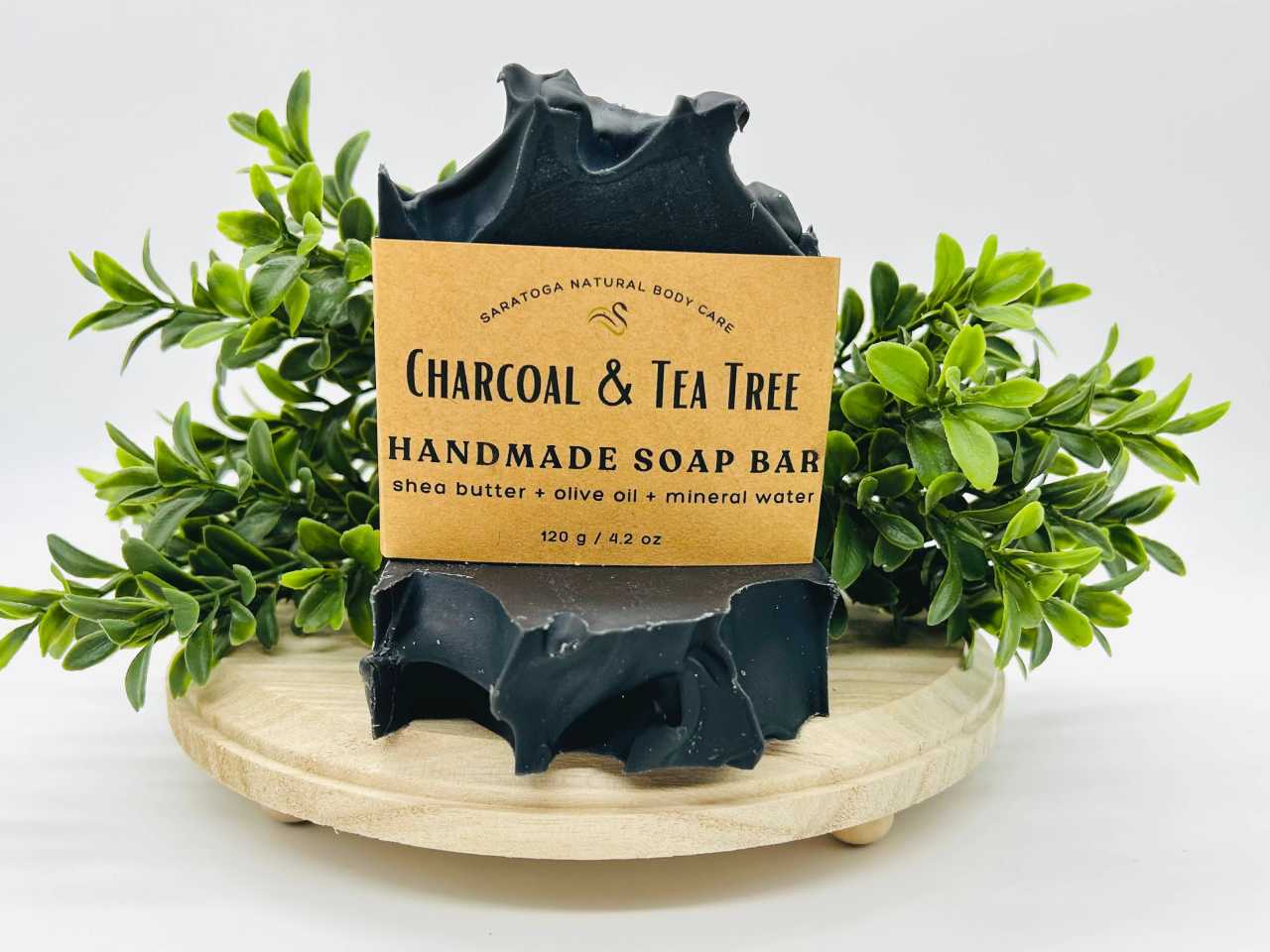 Saratoga Natural Body soap for men featuring charcoal and tea tree crafted with shea butter, olive oil, and mineral water. The artisanal soap features a marbled green and beige design, placed on a wooden soap dish with fresh greenery in the background, emphasizing a natural and eco-friendly skincare product.