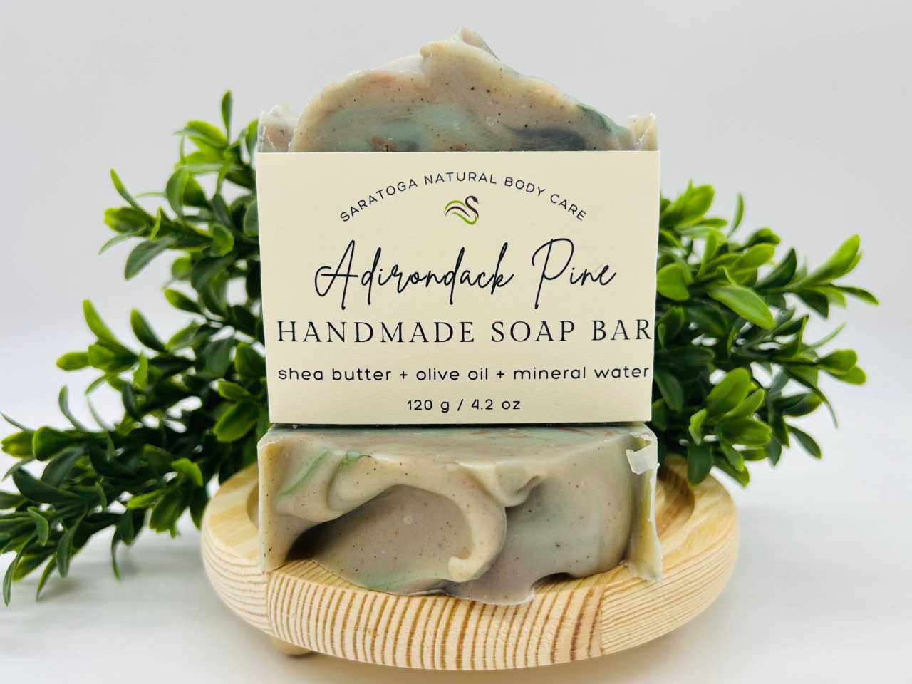 Saratoga Natural Body Care Adirondack Pine handmade soap bar, crafted with shea butter, olive oil, and mineral water. The artisanal soap features a marbled green and beige design, placed on a wooden soap dish with fresh greenery in the background, emphasizing a natural and eco-friendly skincare product.