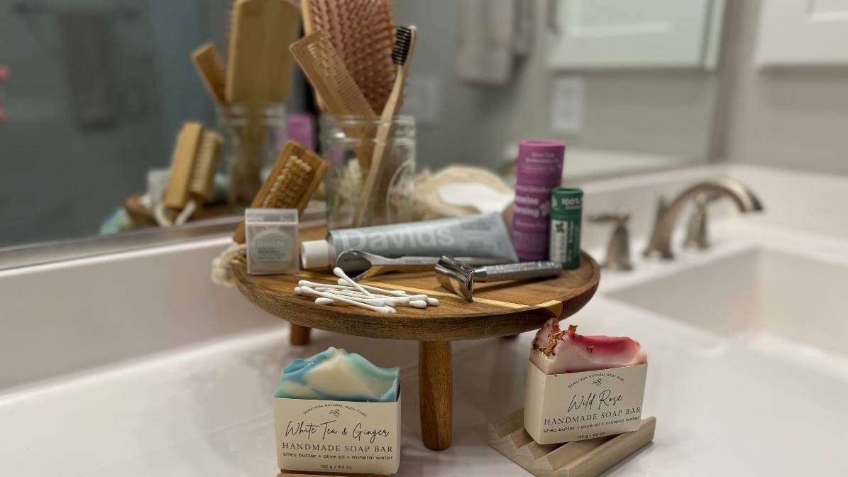 
Eco-friendly bathroom setup with wooden bathroom accessories including bamboo toothbrushes in glass jar, safety razor, cotton swabs, and natural toiletries on wooden display stand. Features handmade artisanal soap bars (White Tea & Ginger and Wild Rose) with shea butter on stylish wooden soap dish. Sustainable zero-waste bathroom essentials with plastic-free alternatives displayed on white sink.
