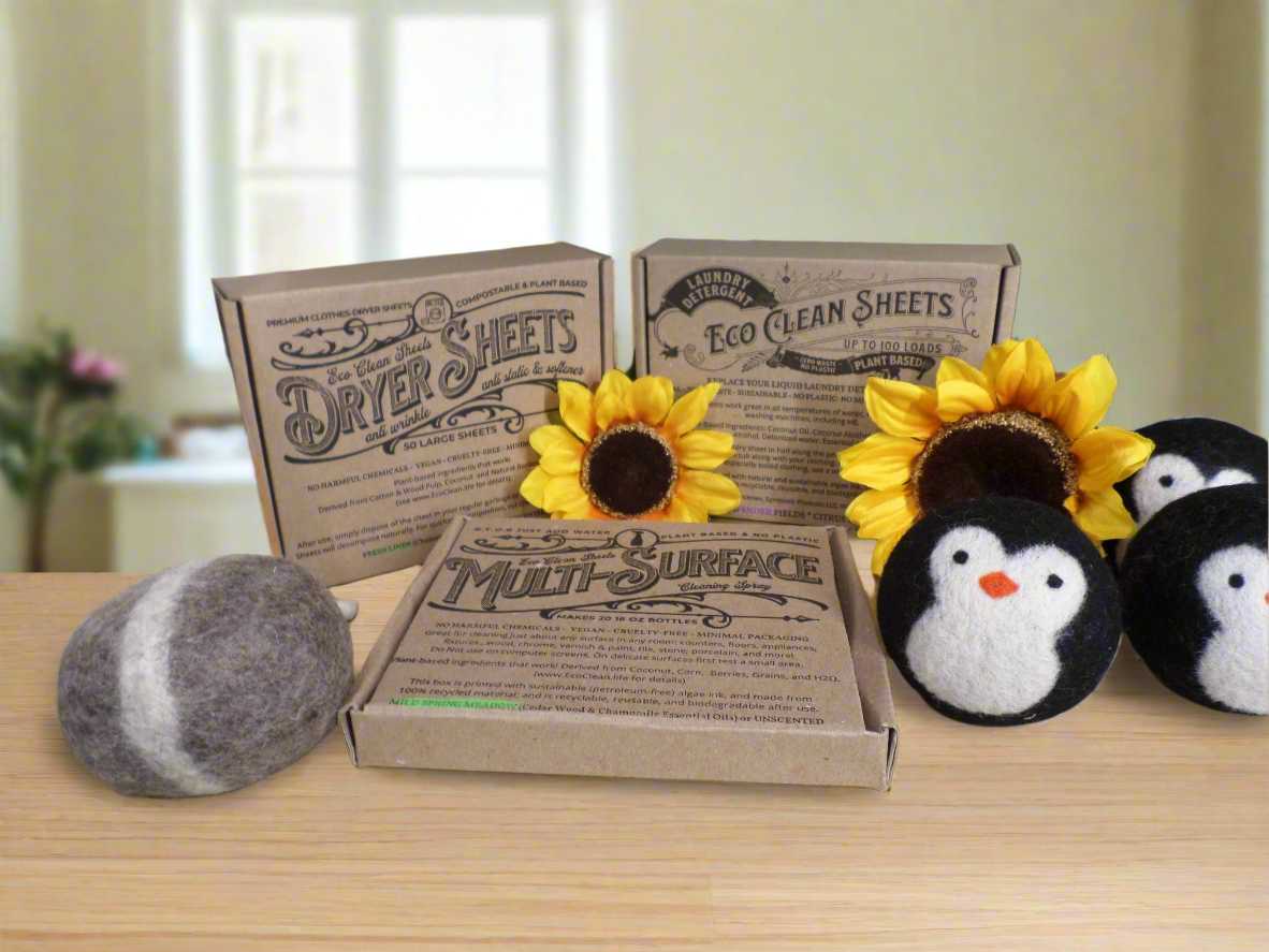 Eco-friendly cleaning products display with kraft paper packaging including Eco Clean Dryer Sheets, Eco Clean Sheets laundry detergent, and Multi-Surface cleaner alongside decorative wool dryer ball, sunflowers, and penguin plush toys on wooden surface. Sustainable home products for chemical-free cleaning.