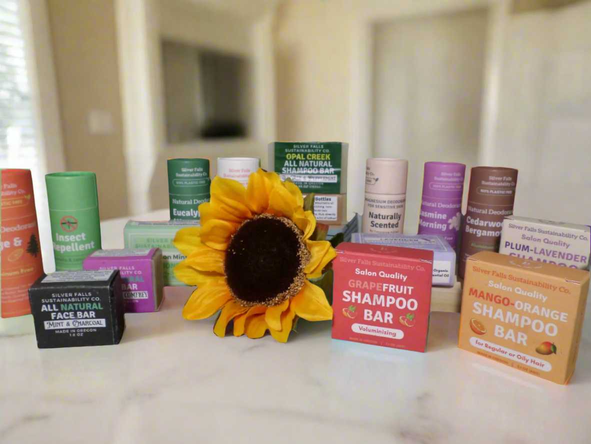 Silver Falls Sustainability Co. zero-waste personal care collection featuring natural shampoo bars in Grapefruit, Mango-Orange, and Plum-Lavender scents, all-natural face bar with mint and charcoal, various natural deodorants in eucalyptus and jasmine scents, insect repellent, and plastic-free packaging displayed with a vibrant sunflower on marble countertop. Eco-friendly bath and body products made in Oregon