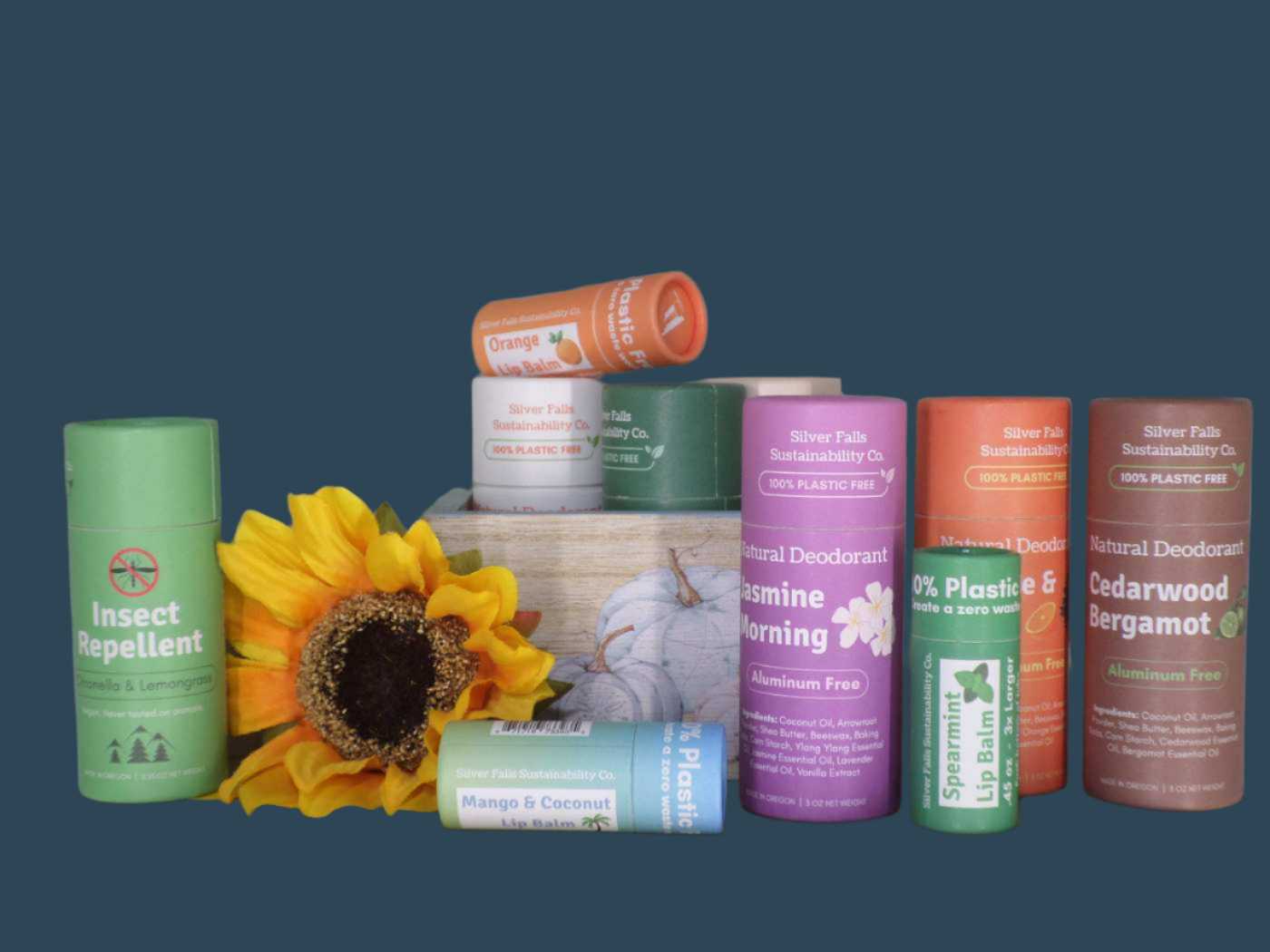 Eco-friendly personal care products from Silver Falls Sustainability Co., including plastic-free natural deodorants, lip balms, and insect repellent in colorful biodegradable packaging. Scents featured include Jasmine Morning, Cedarwood Bergamot, Orange, Mango & Coconut, and Spearmint. Displayed with a sunflower and rustic wooden box for a natural aesthetic.